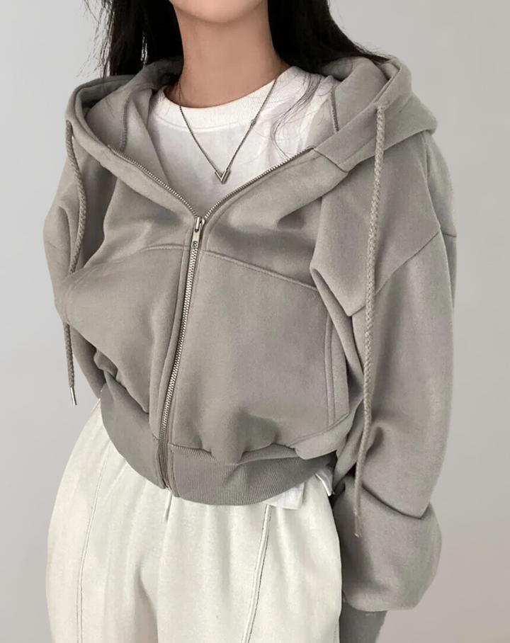 Frontansicht in Grau: Kurze Kapuzenjacke mit geschlossenem Reißverschluss, Kordeln und breitem Rippbündchen, kombiniert mit weißem Shirt und heller Hose. 4