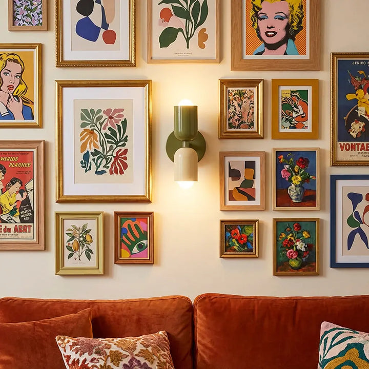 Wandlampe mit olivgrünem und cremefarbenem Schirm mitten in einer Bildergalerie mit Goldrahmen, darunter ein rostrotes Samtsofa mit Kissen. 6