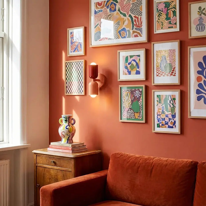 Wandlampe mit dunkelrotem oberem und lachsfarbenem unterem Schirm an einer orangefarbenen Wand zwischen bunten Bilderrahmen über einem Sofa. 5