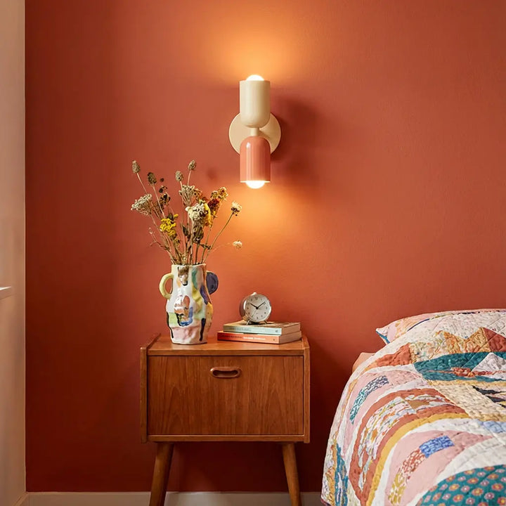 Leuchtende Wandlampe in Creme und Lachsrosa an einer terrakottafarbenen Wand über einem Holz-Nachttisch mit bunter Vase und Trockenblumen. 1