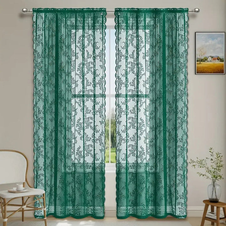 Zwei grüne Spitzenpaneele mit Blumenmuster hängen an einer Stange vor einem Fenster, daneben ein Stuhl und ein Beistelltisch. 5