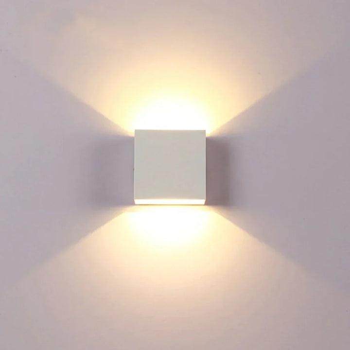 Frontansicht einer weißen Würfelleuchte, die an einer hellen Wand warmes Licht nach oben und unten ausstrahlt. 17