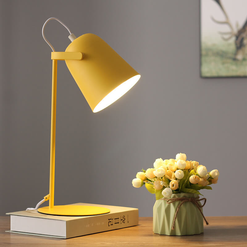 Leuchtende gelbe Tischlampe auf einem Buch vor grauer Wand, daneben eine grüne Vase mit Röschen und ein Bild im Hintergrund. 7