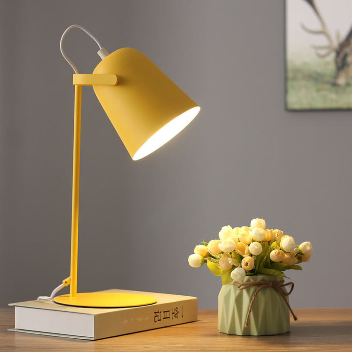 Leuchtende gelbe Tischlampe auf einem Buch vor grauer Wand, daneben eine grüne Vase mit Röschen und ein Bild im Hintergrund. 7