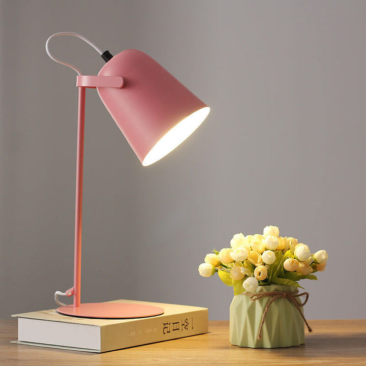 Rosafarbene Tischlampe mit eingeschaltetem Licht auf einem Buch, daneben eine Vase mit hellen Röschen auf einem Holztisch. 8