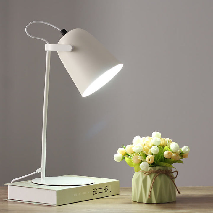 Weiße Tischlampe mit hellem Licht auf einem Buch, daneben eine grüne Vase mit Röschen auf einem Holztisch. 11