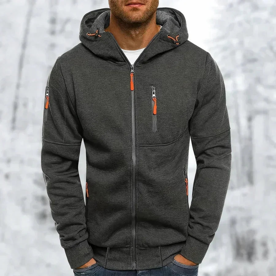 Mann im dunkelgrauen Kapuzenpullover mit orangefarbenen Reißverschlussziehern, Rippenbündchen und Kapuze mit Zugband vor verschneitem Hintergrund. 9