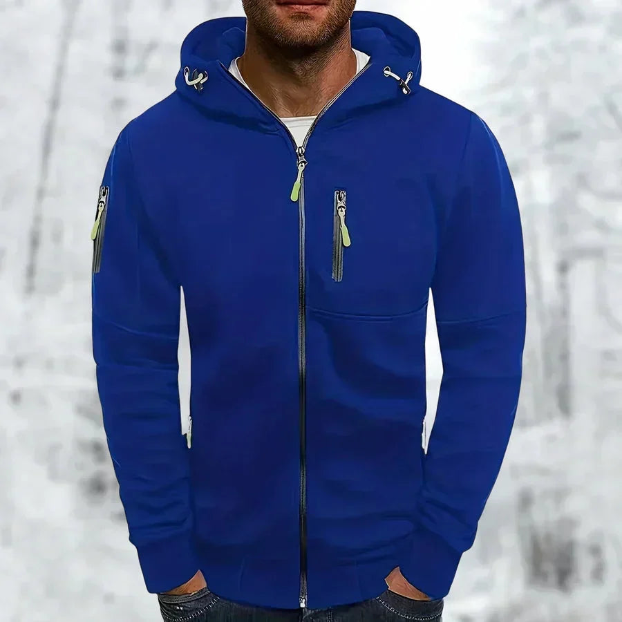 Mann trägt den Kapuzenpullover in Königsblau mit neongrünen Reißverschlussziehern, im Hintergrund eine verschneite Winterlandschaft. 1