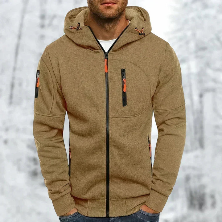 Kapuzenpullover in sandigem Khaki-Braun mit schwarzem Frontreißverschluss und orangefarbenen Zugern, getragen von einem Mann vor Schneelandschaft. 10