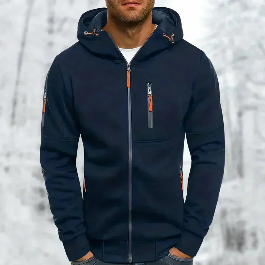 Mann trägt den Kapuzenpullover in Marineblau mit orangefarbenen Reißverschlussziehern und Zugbändern an der Kapuze vor Winterkulisse. 5