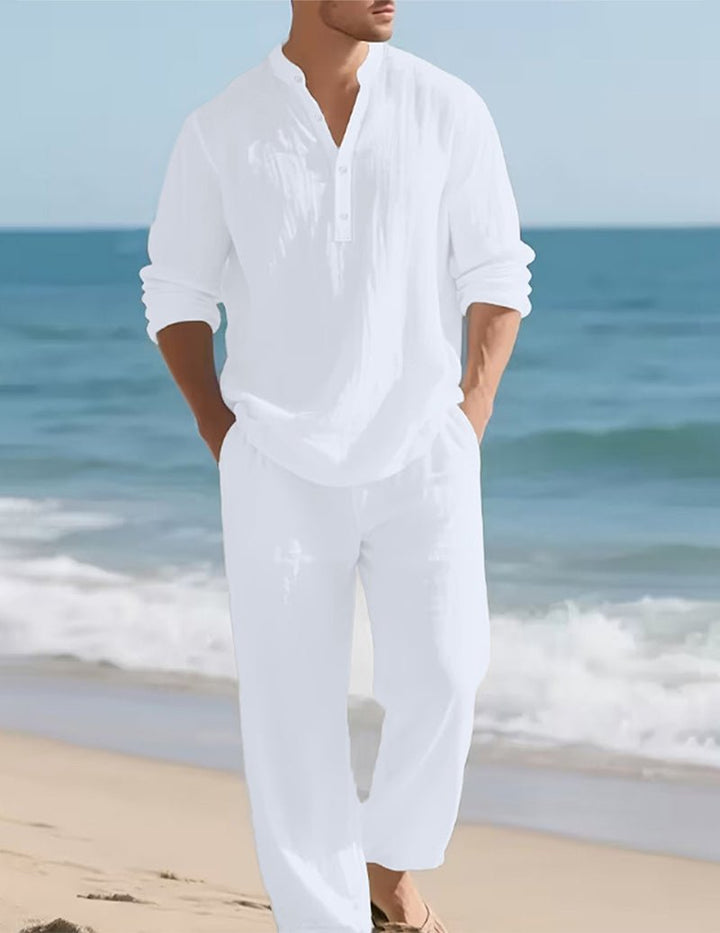 Model im weißen Set mit Knopfleiste und Stehkragen, Hände in den Hosentaschen, am Strand vor dem Meer. 2