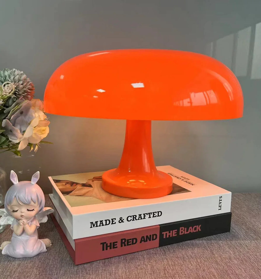 Orange Pilzlampe auf einem Stapel Bücher, daneben ein Blumenstrauß und eine kleine Engelsfigur. 7