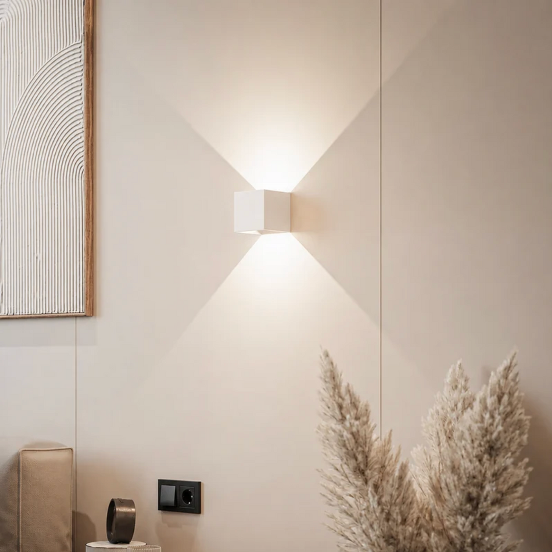 Weiße Würfelleuchte an einer beigen Wand mit Wandbild, Steckdose und Pampasgras, Licht nach oben und unten verteilt. 12
