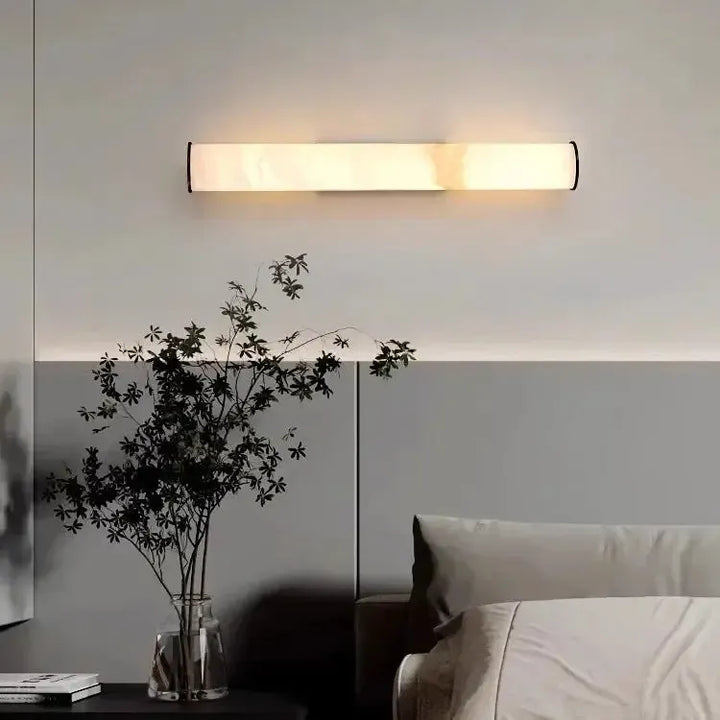 Waagerecht an der Wand montierte Marmor-Wandleuchte über einem Bett, daneben eine Glasvase mit dunklen Zweigen auf einem Nachttisch. 16