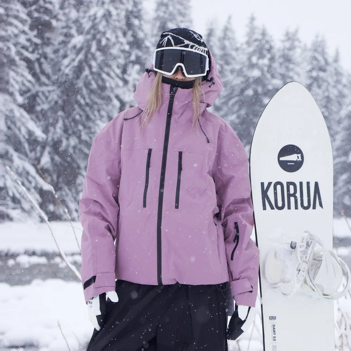 Person in rosafarbener Skijacke mit Kapuze und schwarzen Reißverschlüssen steht im Schneefall neben einem aufrecht gehaltenen weißen Snowboard. 3