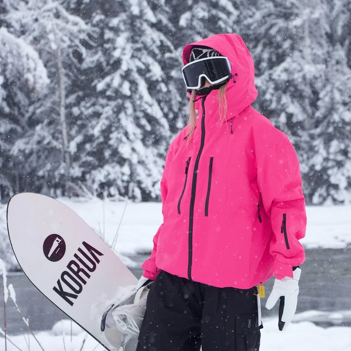 Person in leuchtend pinker Skijacke mit aufgesetzter Kapuze trägt ein Snowboard unter dem Arm vor verschneiten Tannen. 5