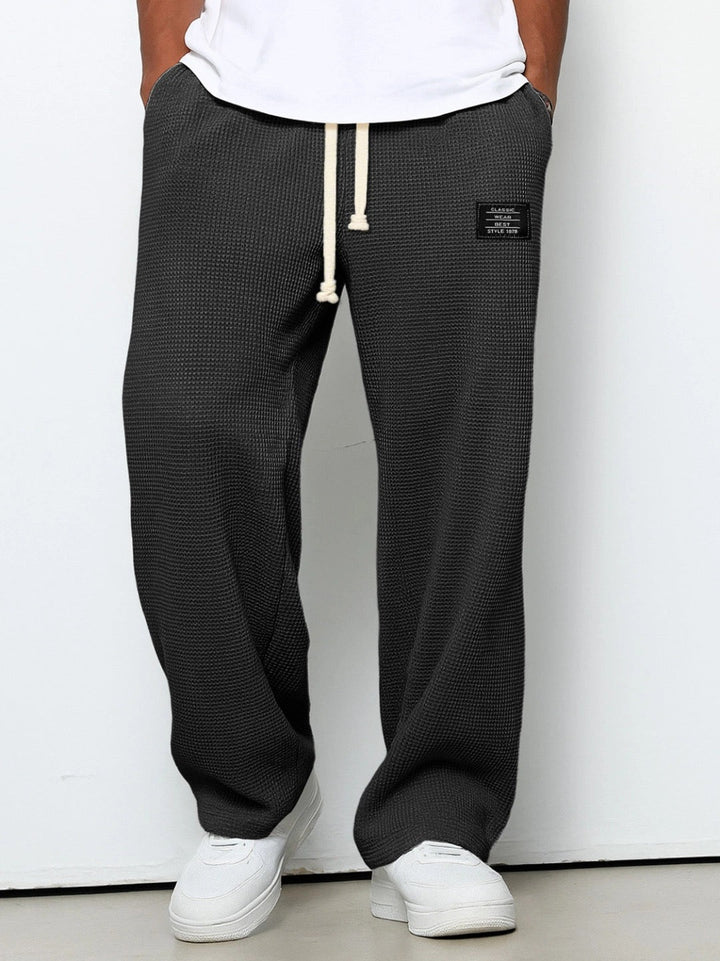 Frontansicht der Waffelstrick-Hose in Anthrazit, getragen von einem Model mit weißem T-Shirt und weißen Sneakern, Hände in den Taschen. 1