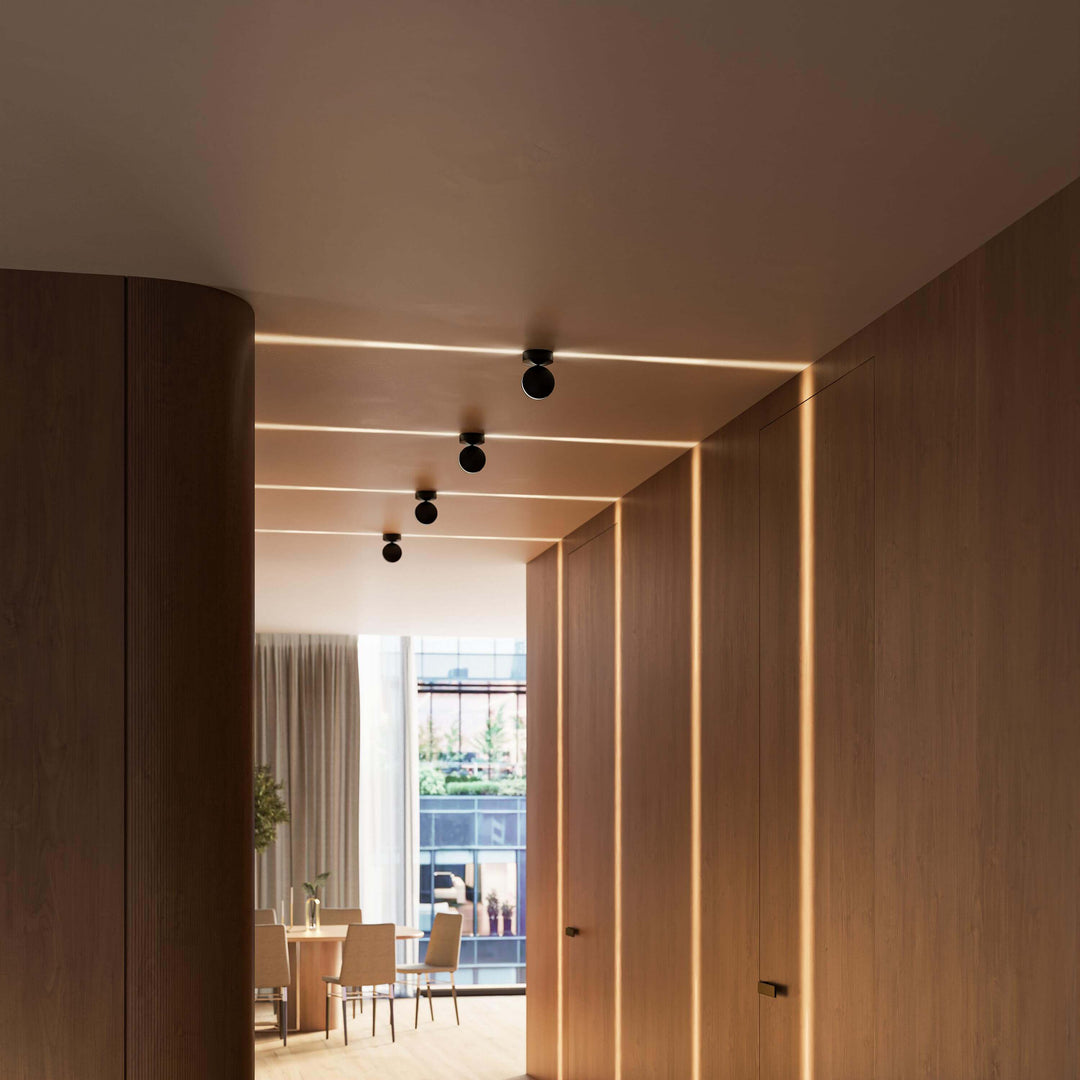 Blick auf vier schwarze Deckenleuchten über einem Holzflur, ihre Lichtbänder laufen quer über die Decke zu den Wandpaneelen. 15