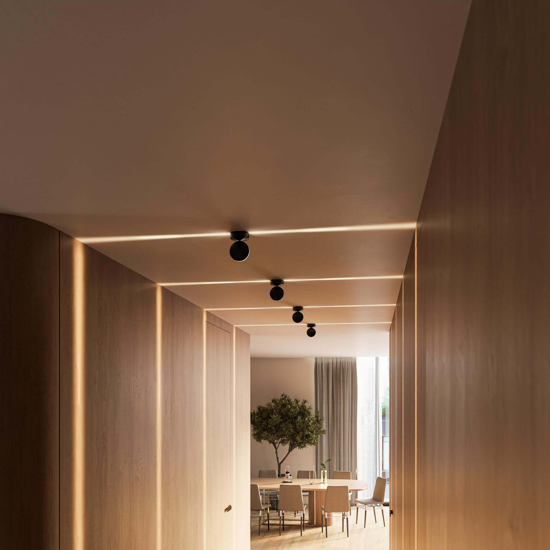 Schräge Ansicht eines Holzflurs mit vier schwarzen Deckenleuchten, deren Licht als feine Linien über Decke und Wände fällt. 17