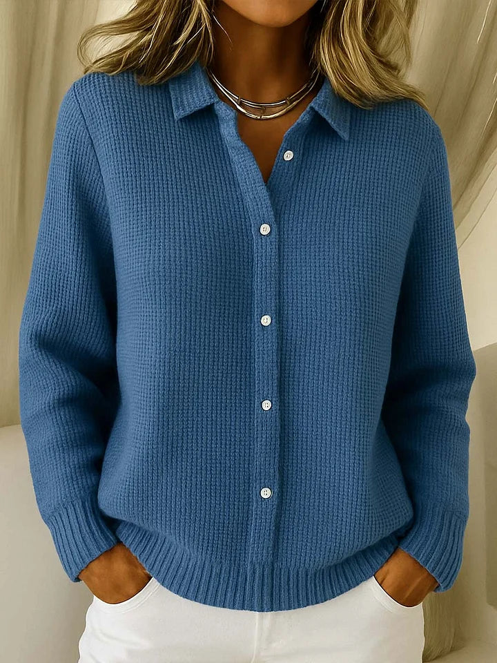 Der Strickpullover in mittlerem Blau am Model, Vorderansicht mit Kragen, Knopfleiste und gerippten Bündchen, kombiniert mit weißer Hose. 4