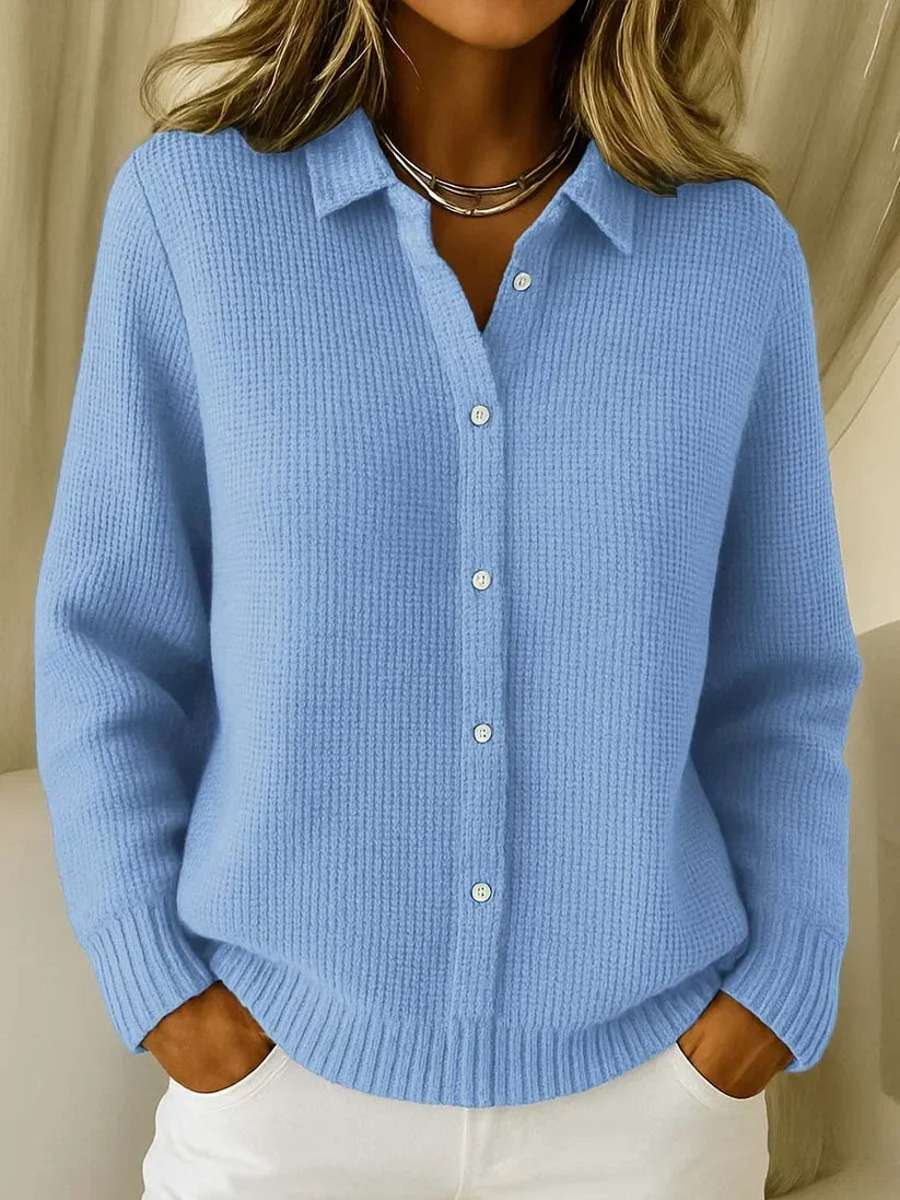 Frau trägt den strukturierten Strickpullover in Hellblau mit Kragen und Knopfleiste, die Hände in den Taschen einer weißen Hose. 1