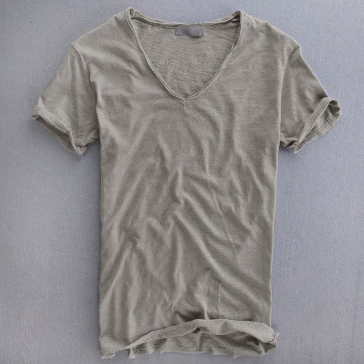 Kurzarm-T-Shirt mit tiefem V-Ausschnitt in Graubeige, flach auf grauem Untergrund ausgebreitet, Ärmelsäume nach oben umgeschlagen. 1