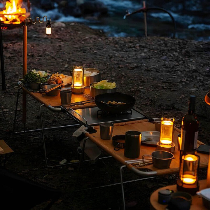 Mehrere leuchtende Lampen auf Campingtischen mit Kochgeschirr und Essen, abends im Freien am Feuer. 4