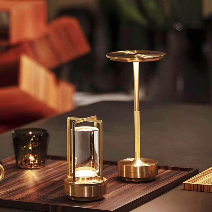 Goldene Lampe mit Glaszylinder auf einem Holztablett, daneben eine schlanke goldene Lampe mit rundem Schirm. 7