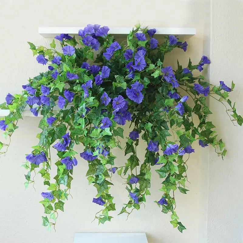 Violettblaue Kunstblüten mit langen hellgrünen Blattranken, die von einem weißen Wandregal über eine cremefarbene Wand herabhängen. 4