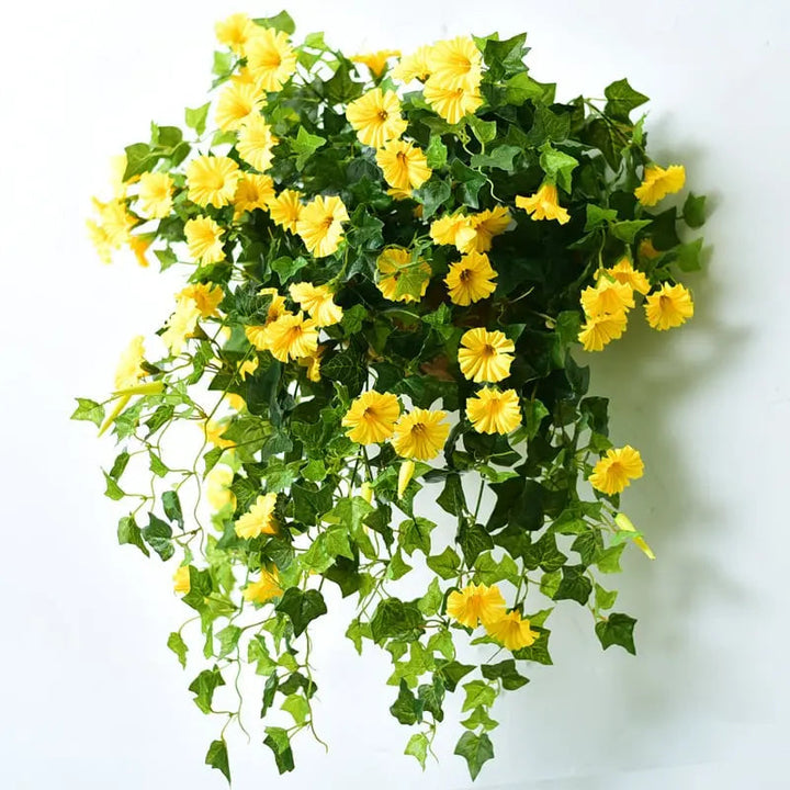 Gelbe Kunstblüten mit grünen Efeublättern als hängendes Arrangement vor weißer Wand, von vorn fotografiert, mit weichem Schattenwurf. 5