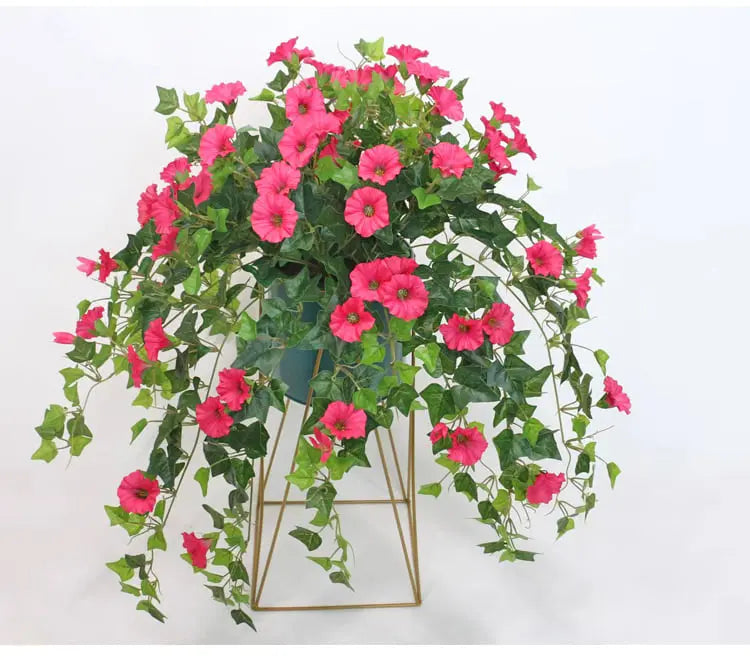 Pinkfarbene Kunstblumen mit überhängenden Ranken in einem dunklen Topf auf einem goldfarbenen Drahtgestell vor weißem Hintergrund. 3