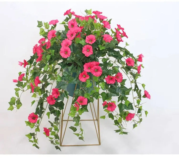 Pinkfarbene Kunstblumen mit überhängenden Ranken in einem dunklen Topf auf einem goldfarbenen Drahtgestell vor weißem Hintergrund. 3