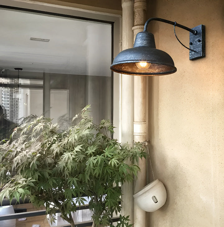 Wandlampe mit patinierter Oberfläche leuchtet an einer cremefarbenen Hauswand neben einem Fallrohr, davor ein Ahornbaum und ein weißer Hängetopf. 3