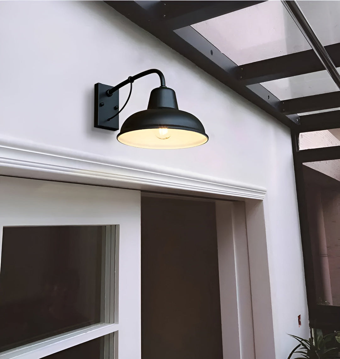 Schwarze Wandlampe mit leuchtender Glühbirne an einer weißen Fassade über einer Tür, darüber ein Vordach mit dunklem Gestell. 4