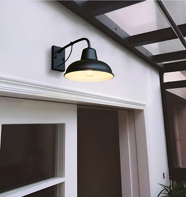 Schwarze Wandlampe mit leuchtender Glühbirne an einer weißen Fassade über einer Tür, darüber ein Vordach mit dunklem Gestell. 4