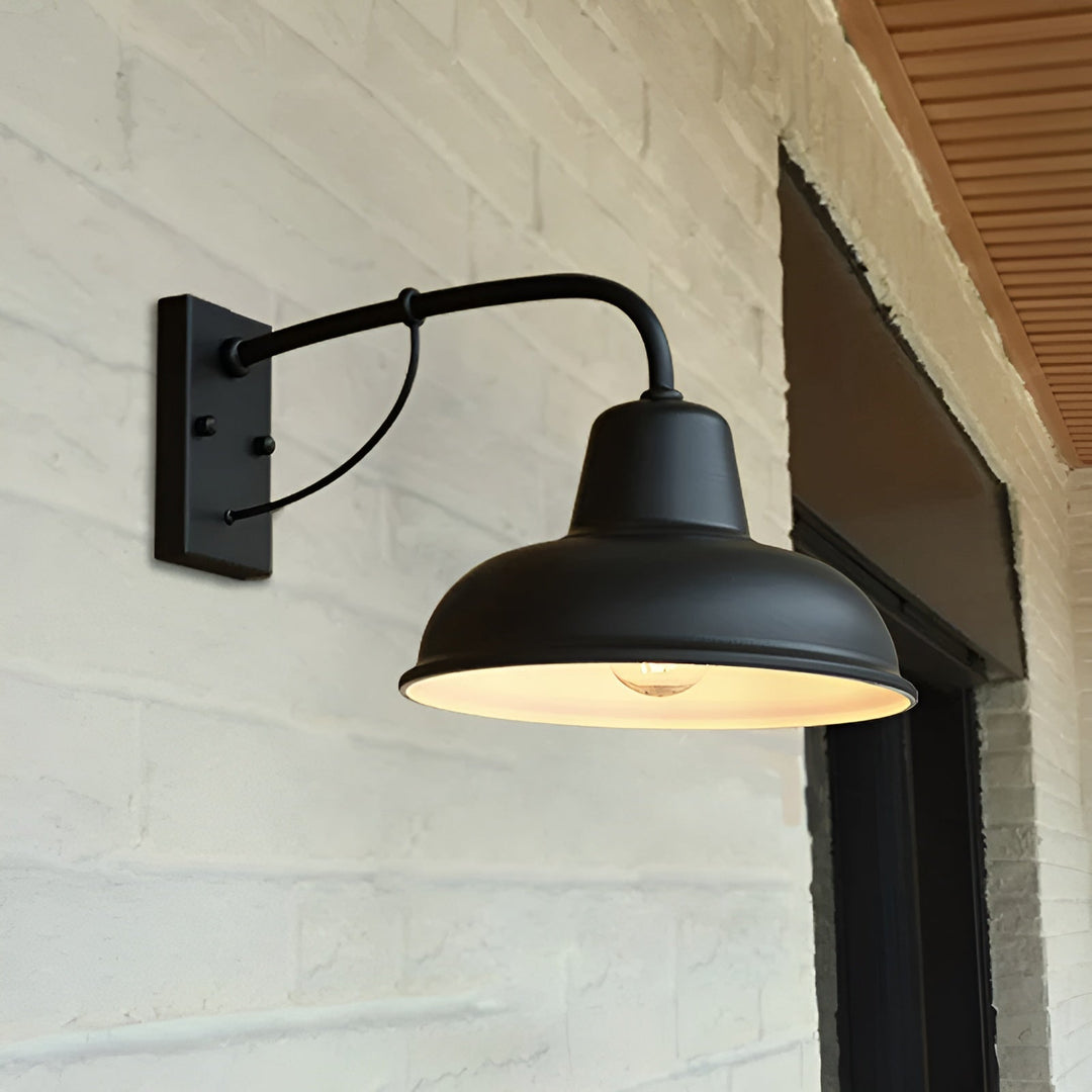 Schwarze Wandlampe mit gebogenem Arm, an einer hell gestrichenen Backsteinwand neben einer Türöffnung montiert, der Schirm ist innen hell und beleuchtet. 5