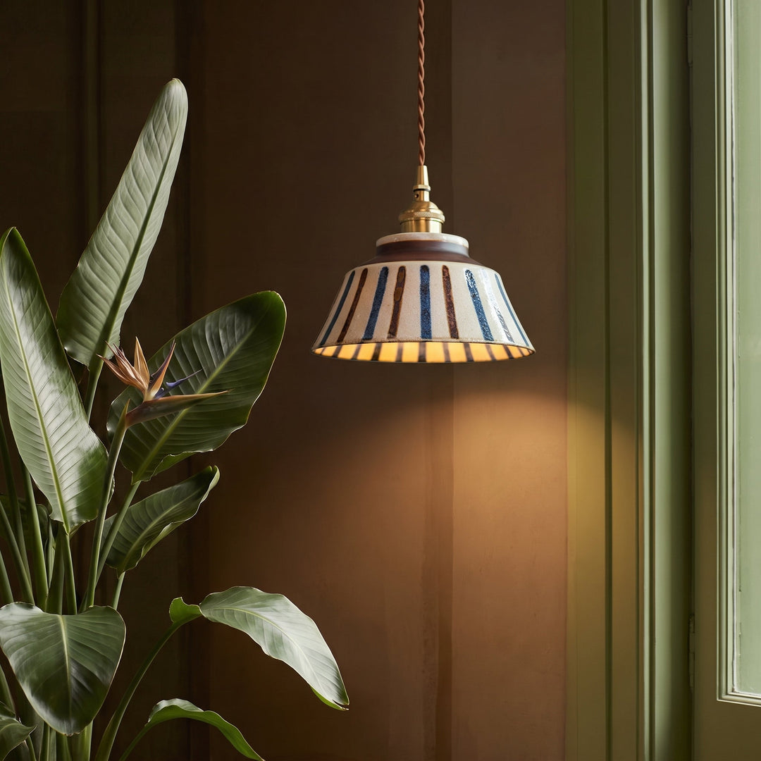 Gestreifte Keramik-Pendelleuchte neben einer Paradiesvogelblume vor brauner Wand, die warmes Licht an die Wand wirft. 4