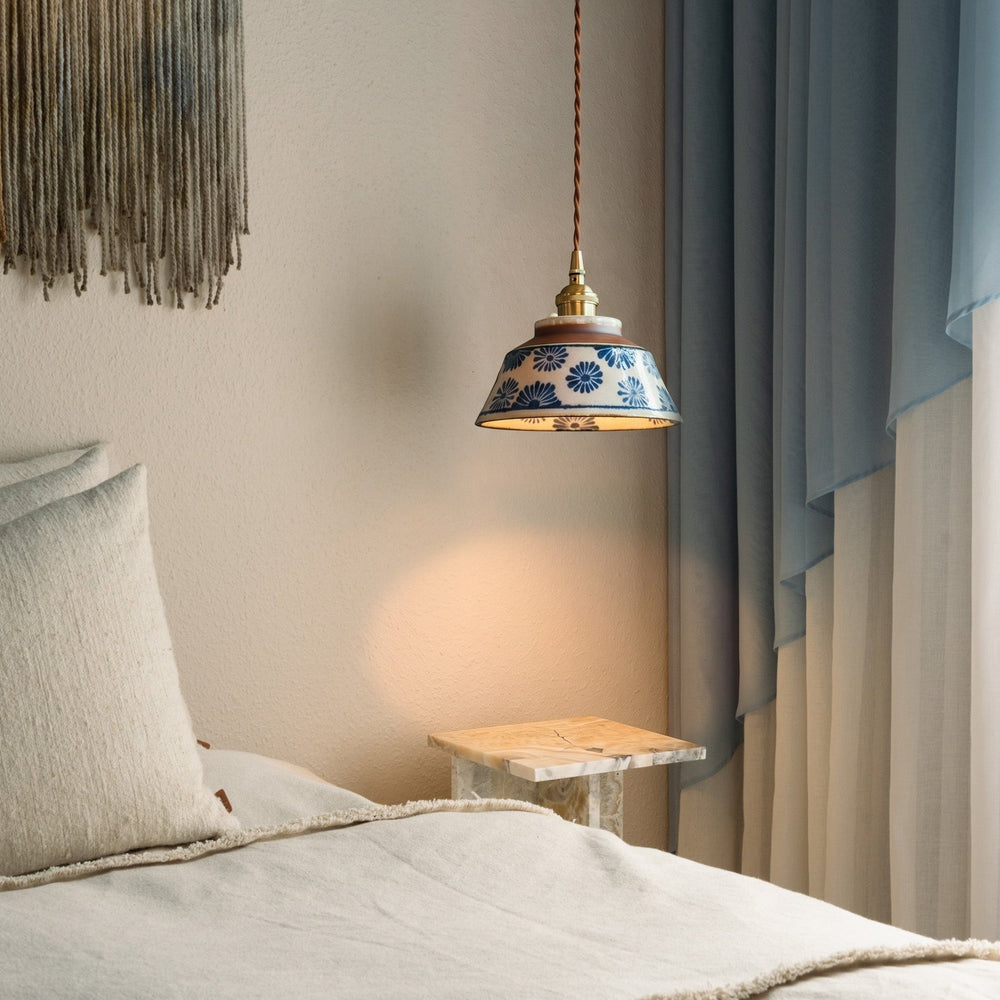 Pendelleuchte mit blauem Blumenmuster über einem Marmor-Nachttisch neben Bett, Kissen und hellblauem Vorhang im warmen Licht. 2