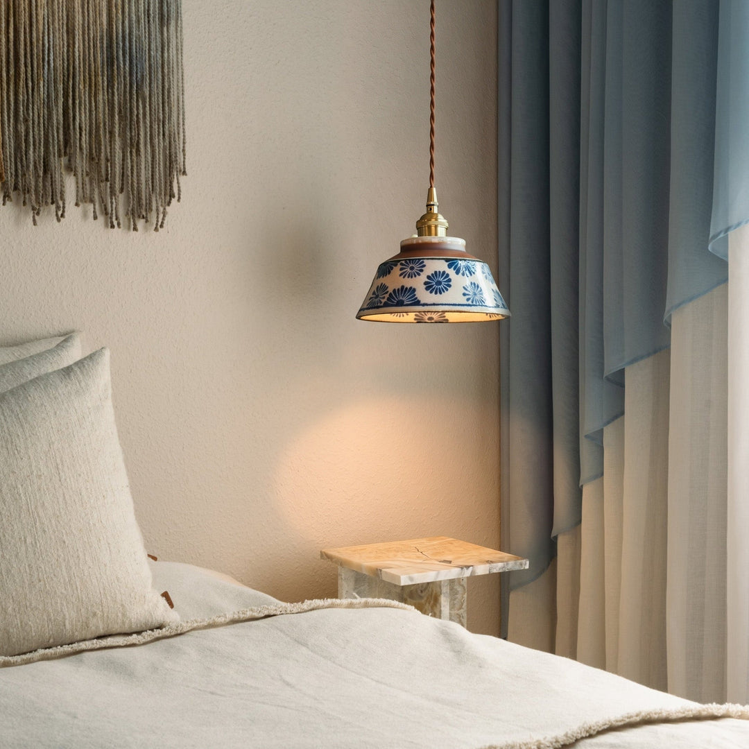 Pendelleuchte mit blauem Blumenmuster über einem Marmor-Nachttisch neben Bett, Kissen und hellblauem Vorhang im warmen Licht. 2