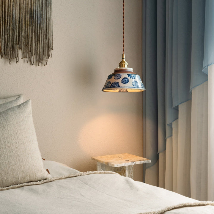 Pendelleuchte mit blauem Blumenmuster über einem Marmor-Nachttisch neben Bett, Kissen und hellblauem Vorhang im warmen Licht. 2