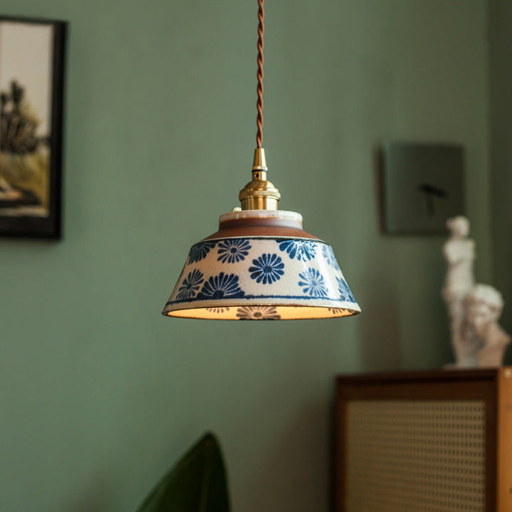 Leuchtende Keramikleuchte mit blauem Blumenmuster, Messingfassung und braunem Stoffkabel vor einer grünen Wand mit Bild und Statue. 1