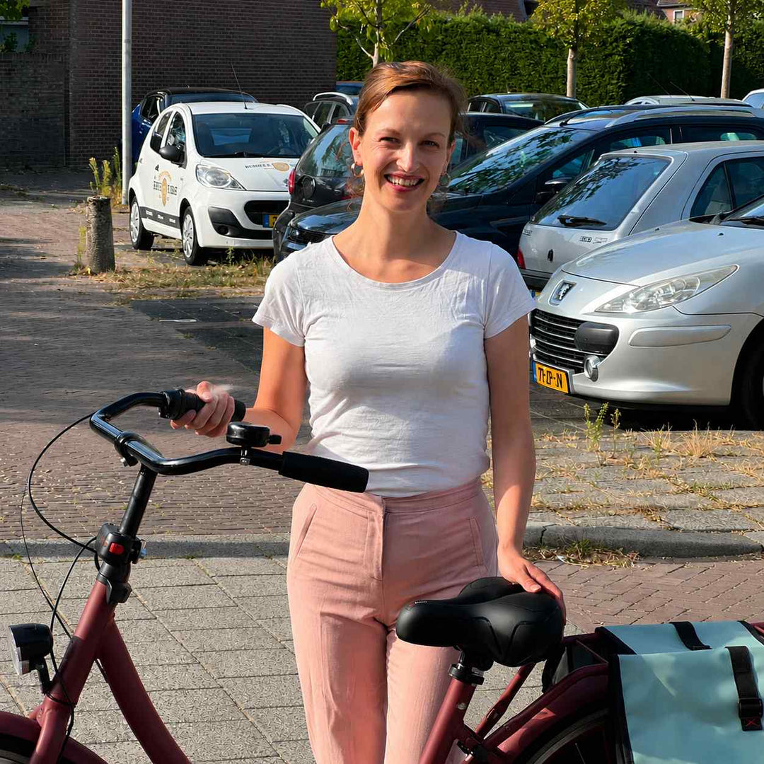 Lächelnde Frau steht neben einem roten Fahrrad und legt die Hand auf den schwarzen Komfortsattel, daneben eine Satteltasche. 7