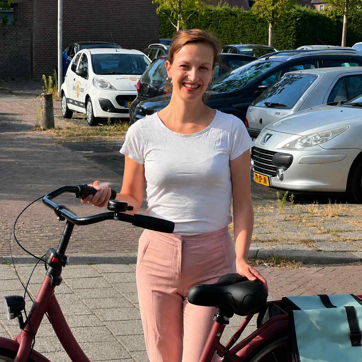 Lächelnde Frau steht neben einem roten Fahrrad und legt die Hand auf den schwarzen Komfortsattel, daneben eine Satteltasche. 7