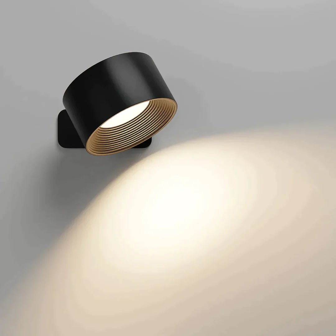 Schwarze Wandlampe mit goldfarbenem, gerilltem Innenring, schräg gestellt, strahlt warmweißes Licht auf die Wand. 2