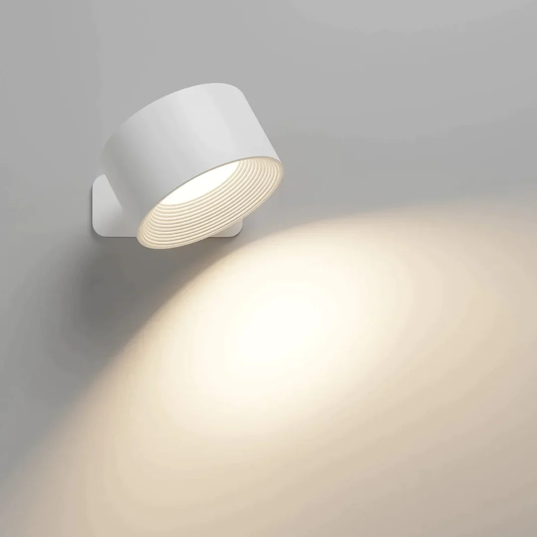 Weiße zylindrische Wandlampe an heller Wand, schräg nach unten geneigt, wirft einen warmen, weichen Lichtkegel. 1