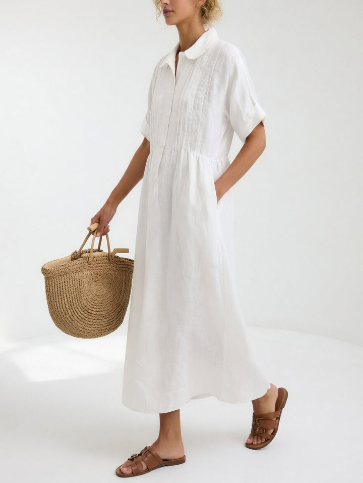 Seitliche Ansicht des weißen Maxi-Hemdkleids mit Biesen an der Brust, getragen mit Strohtasche und braunen Sandalen, eine Hand in der Tasche. 1