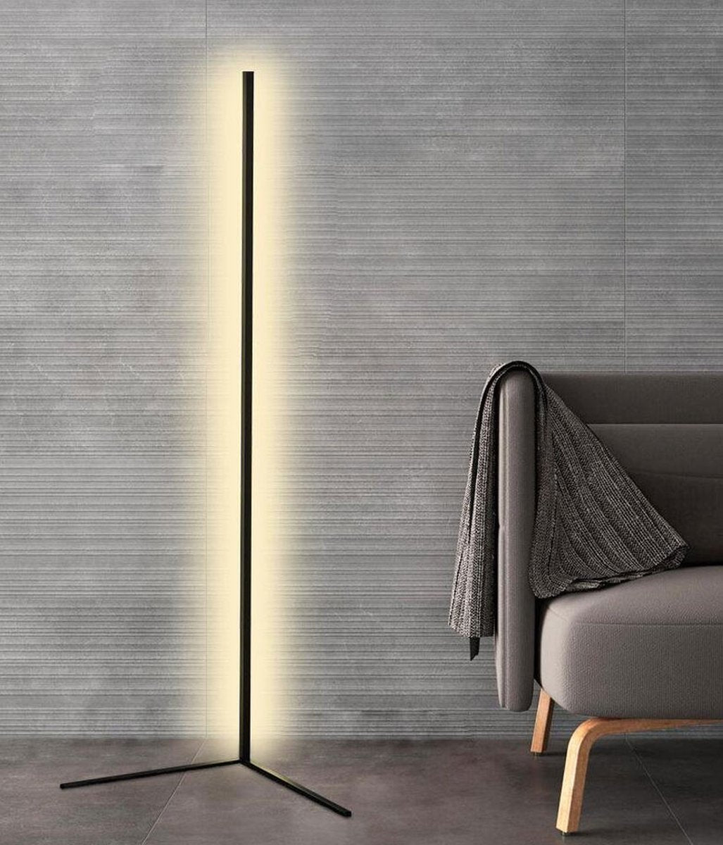 Die schwarze Stehlampe mit L-förmigem Fuß leuchtet vor einer grauen Strukturwand neben einem Sofa mit Decke 8