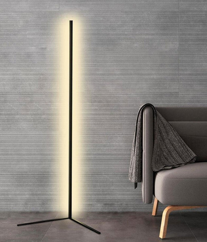 Die schwarze Stehlampe mit L-förmigem Fuß leuchtet vor einer grauen Strukturwand neben einem Sofa mit Decke 8