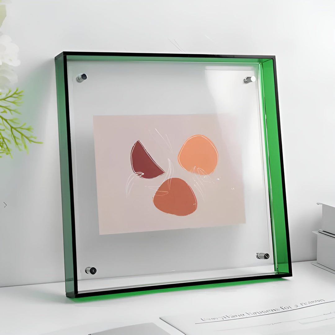 Quadratischer Acrylrahmen mit grünem Rand und sichtbaren Metallhaltern, der ein abstraktes Motiv in Orange- und Rottönen zeigt, auf einem weißen Tisch. 21