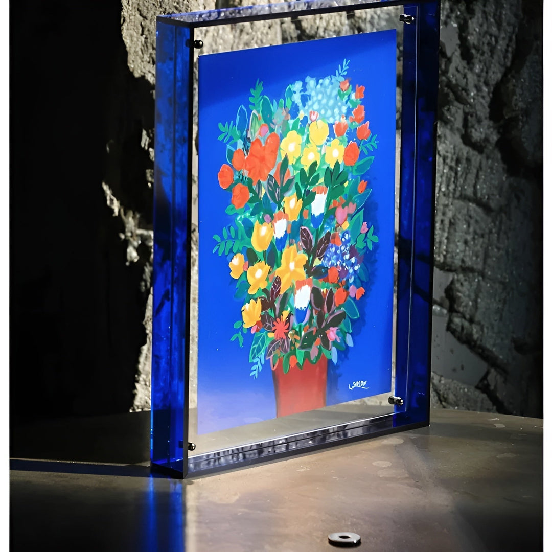 Stehender Rahmen mit tiefblauem Acrylrand vor einer Steinwand, darin ein leuchtendes Blumenstrauß-Bild auf blauem Grund. 4